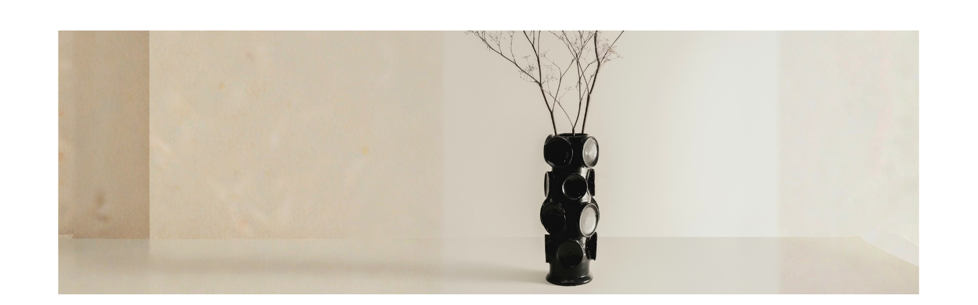 A black geometric vase with dry twigs sits on a minimalist white surface against a neutral beige background.