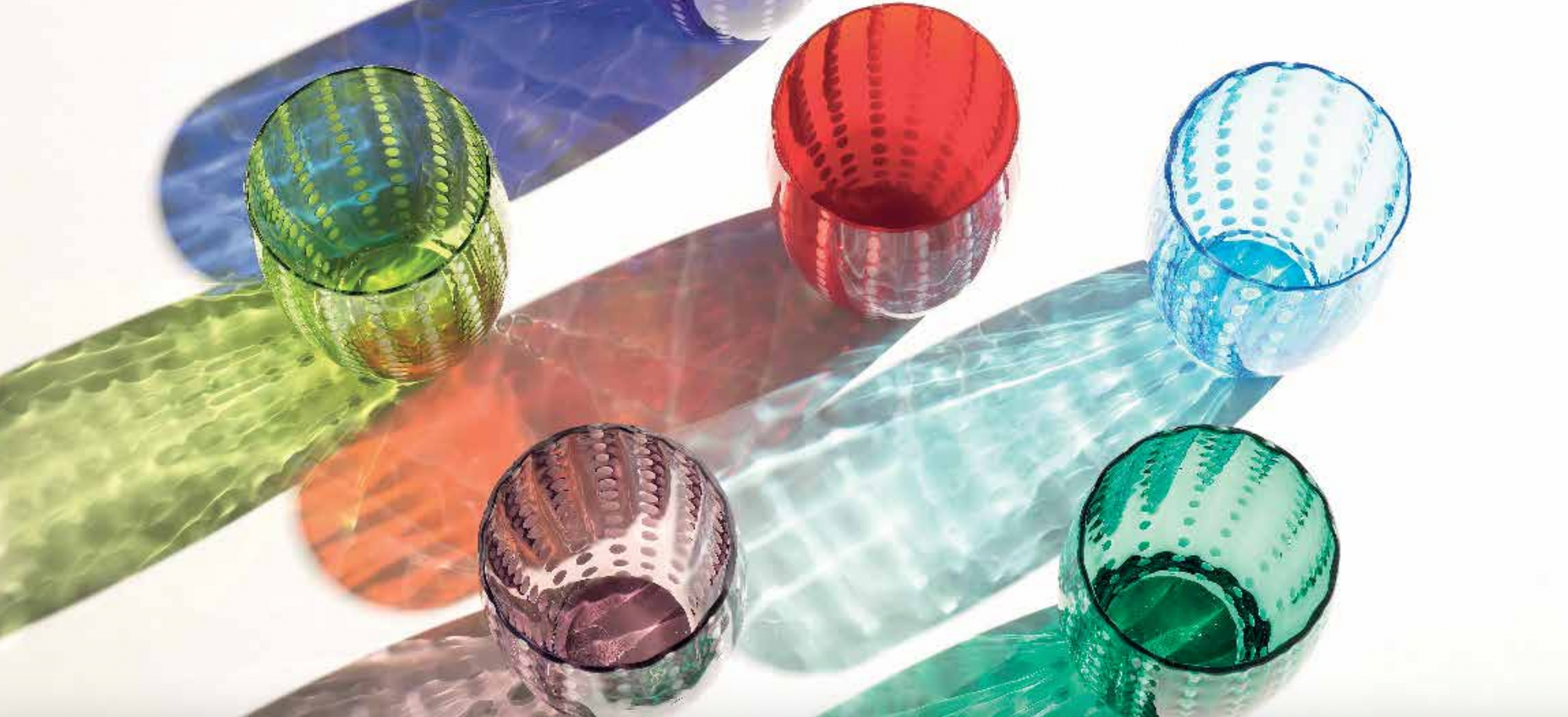 Five colorful, textured glass cups—green, purple, red, blue, and teal—are arranged on a white surface, casting vibrant, patterned shadows in matching hues.