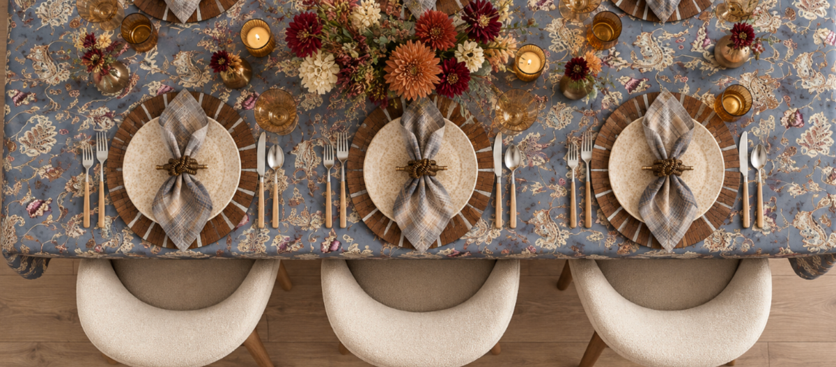 A rustic table setting with a wood-look charger, beige plate, plaid napkin in a woven napkin ring, and two amber glasses on a marble surface. Autumn flowers are partially visible in the corner.