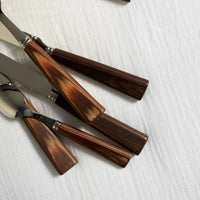 Several pieces from the Sabre Nature Flatware 5 Piece Setting-Light Wood, featuring polished light wood handles and shiny metal blades, are arranged on textured white fabric, showcasing the unique wood grain patterns of each utensil.