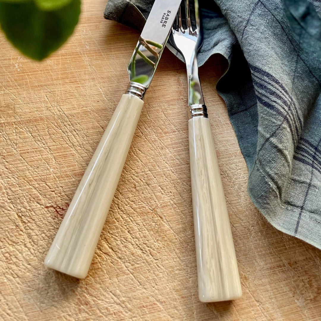 A close-up of the Sabre Nature Flatware 5 Piece Setting - Faux Horn, featuring cream-colored handles, made in France, resting on a wooden surface beside a blue and gray plaid napkin.