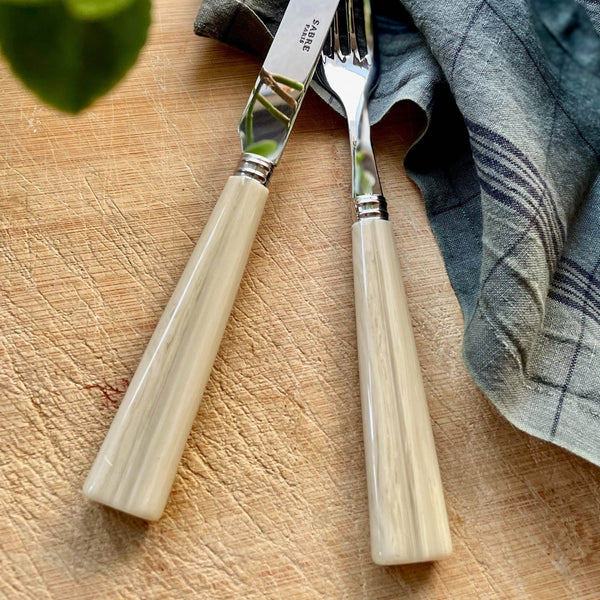A close-up of the Sabre Nature Flatware 5 Piece Setting - Faux Horn, featuring cream-colored handles, made in France, resting on a wooden surface beside a blue and gray plaid napkin.