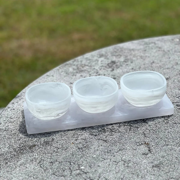 Three small, round bowls from the BT- Shalom Dawn Resin Swirl 3 Dip Bowl Set in white, each featuring a subtle swirl design, rest on a matching rectangular tray atop a textured gray outdoor table with grass in the background.