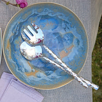 A blue ceramic bowl holds the Michael Wainwright Truro Platinum Salad Server Set, featuring textured platinum handles. A purple napkin rests nearby. The set is dishwasher safe for easy cleaning.