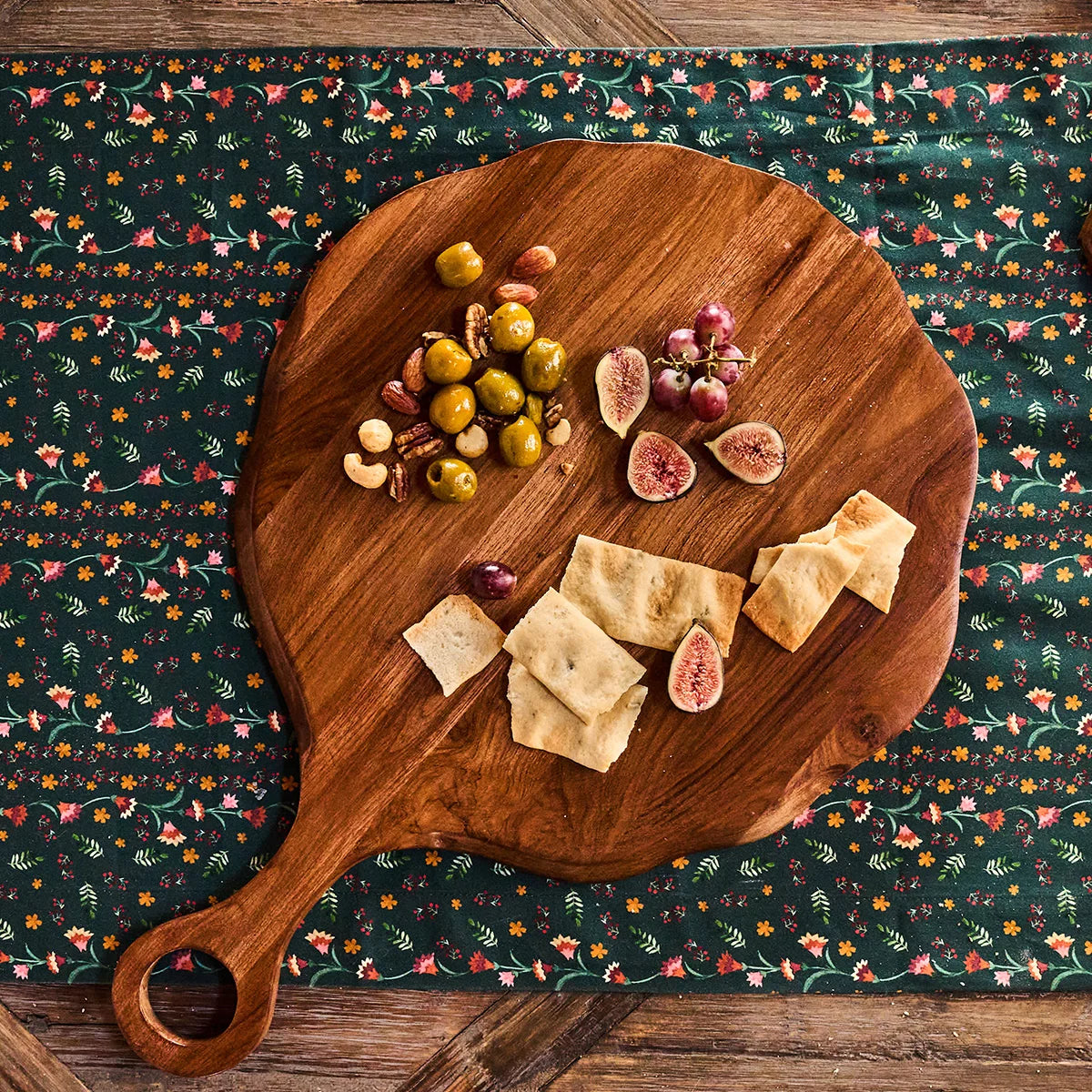 The Juliska Puro Round Serving Board Teak showcases an inviting arrangement of green olives, nuts, figs, grapes, and pieces of flatbread atop a floral-patterned tablecloth—perfect for entertaining with a beautifully presented charcuterie board.