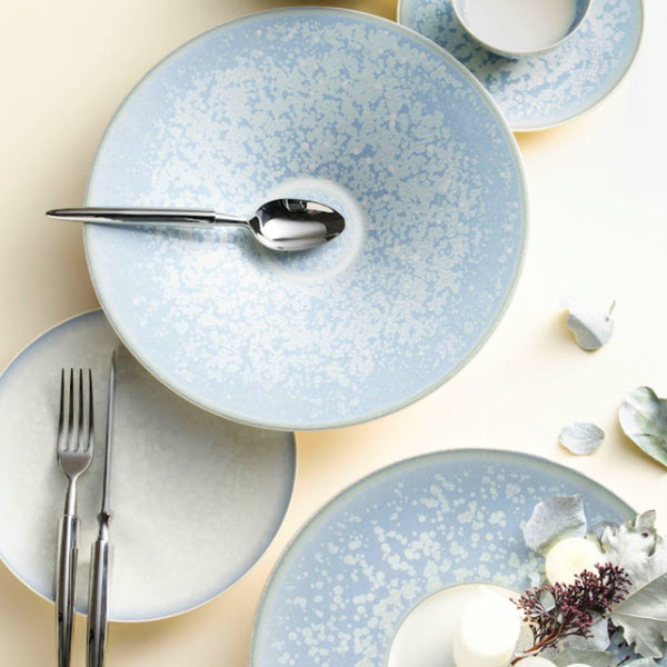 The Song Ocean Dinnerware by Jaune De Chrome features light blue enamel plates with a speckled white pattern, accompanied by a spoon, fork, knife, cup, napkin with white flowers, and candles for a charming French table setting.