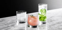 The Richard Brendon Fluted Shot Glass Set of 2 shines on a marble surface, one glass empty and the other with a pink drink, ice, and garnish—elevating any barware collection against a striking black and grey backdrop.