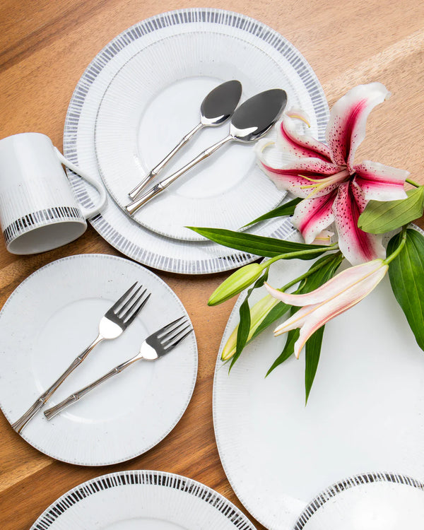 A Ricci Bamboo Flatware 5 Piece Setting with white plates featuring blue rims, a white mug, and pink lilies arranged on a wooden surface.