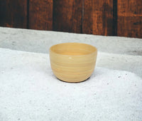 The Albert L Bamboo Classic Serving Bowl Small in Natural rests on a textured white surface with a wooden wall behind it.