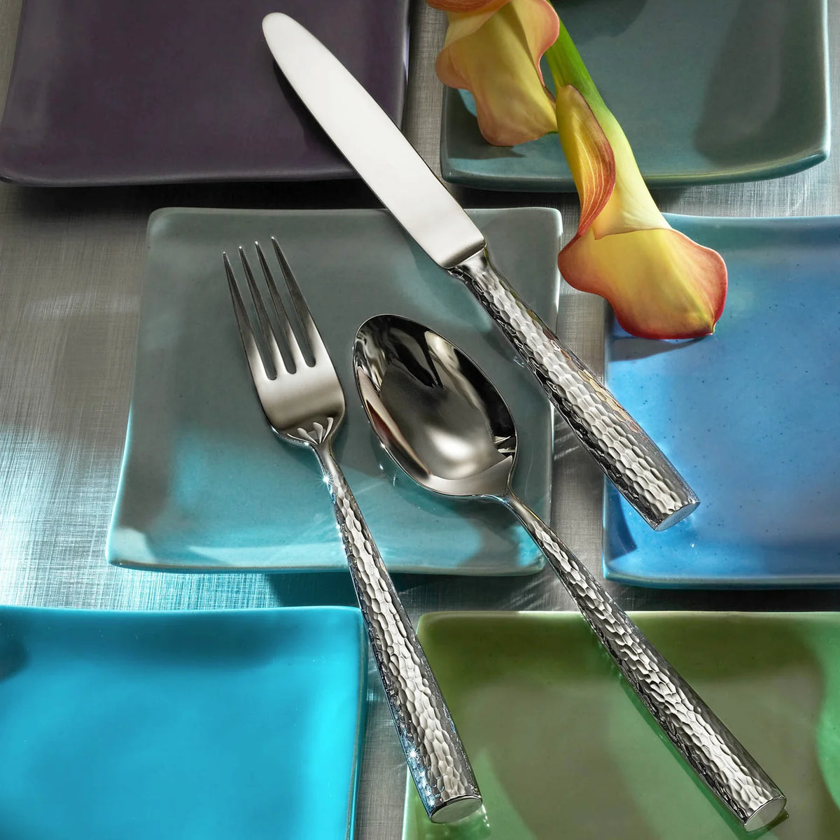 The Ricci Anvil Flatware 5 Piece Setting, featuring hammered texture handles, is displayed on a light blue square plate surrounded by colorful dishes and two orange calla lilies atop a metallic surface.