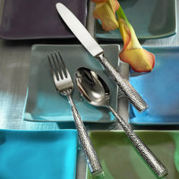 The Ricci Anvil Flatware 5 Piece Setting, featuring hammered texture handles, is displayed on a light blue square plate surrounded by colorful dishes and two orange calla lilies atop a metallic surface.