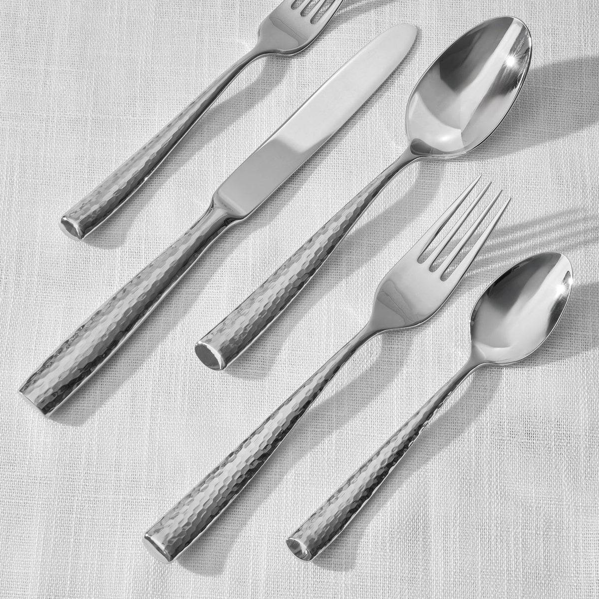 The Ricci Anvil Flatware 5 Piece Setting, featuring a hammered texture, includes two forks, a knife, and two spoons arranged diagonally on a white linen tablecloth.