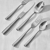 The Ricci Anvil Flatware 5 Piece Setting, featuring a hammered texture, includes two forks, a knife, and two spoons arranged diagonally on a white linen tablecloth.
