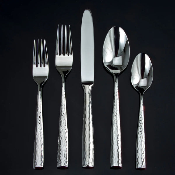 The Ricci Anvil Flatware 5 Piece Setting, crafted from 18/10 stainless steel, includes a salad fork, dinner fork, knife, soup spoon, and teaspoon—each with hammered textured handles and polished surfaces.