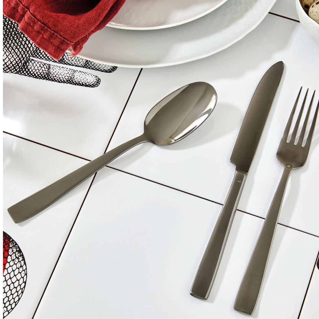 The Sambonet Flat PVD Flatware 5 Piece Setting in Black is arranged on white tiles near plates, a red napkin, and a hand illustration. The sleek stainless steel pieces enhance the modern table setting.