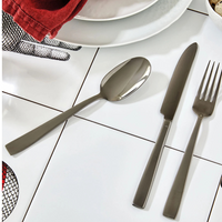 The Sambonet Flat PVD Flatware 5 Piece Setting in Black is arranged on white tiles near plates, a red napkin, and a hand illustration. The sleek stainless steel pieces enhance the modern table setting.
