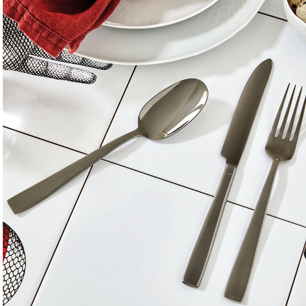 The Sambonet Flat PVD Flatware 5 Piece Setting in Black is arranged on white tiles near plates, a red napkin, and a hand illustration. The sleek stainless steel pieces enhance the modern table setting.