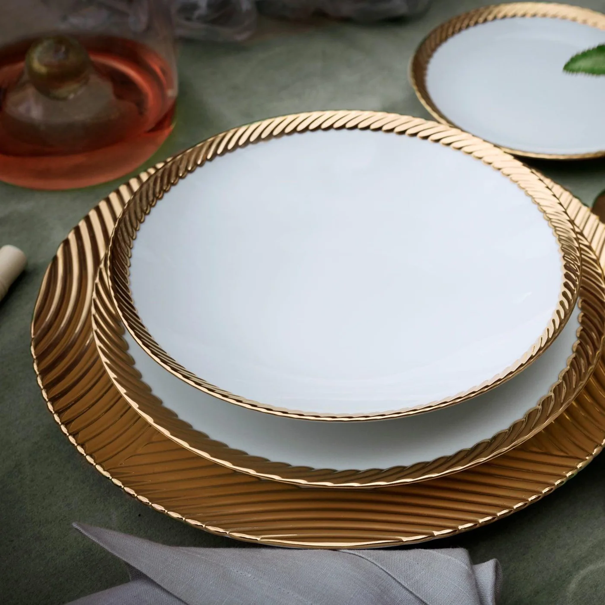 Three stacked Corde Dinnerware Gold plates by L'Objet, featuring white surfaces and gold textured rims, are displayed on a green surface with a glass of pink drink, a cloth napkin, and part of a leaf visible.