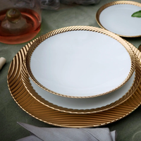 Three stacked Corde Dinnerware Gold plates by L'Objet, featuring white surfaces and gold textured rims, are displayed on a green surface with a glass of pink drink, a cloth napkin, and part of a leaf visible.