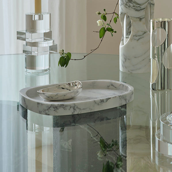 A modern glass table features the Greg Natale Pietra Catchall and a small silver dish, accented by marble and silver vases. A leafy branch and soft reflections bring elegant, organic style to this contemporary arrangement.