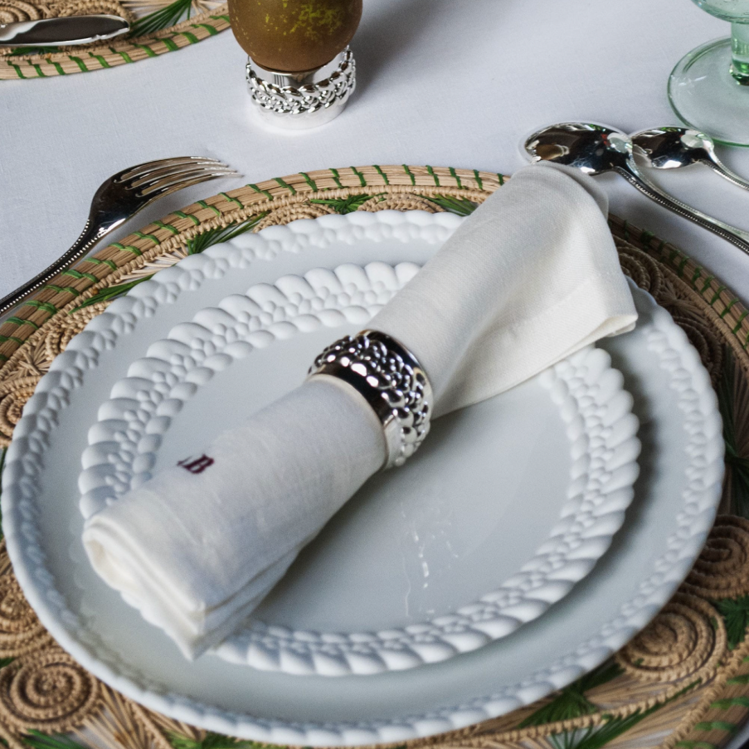 The Christofle Babylone Dinnerware, a white porcelain plate with scalloped edge and beaded detail, is set on a woven placemat with a white napkin in a silver ring, silver cutlery, and a green-tinted glass.