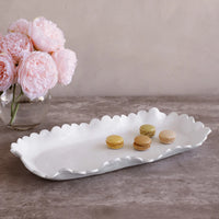 The Beatriz Ball Vida Colette Platter White - Large, featuring wavy edges, displays four colorful macarons on a gray surface beside a glass vase of soft pink peonies.