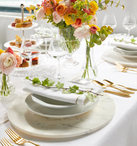 An elegant table setting features Sferra’s Seaton Napkin Flint & Gold Set of 4, gold cutlery, white plates on marble chargers, wine glasses, a floral centerpiece, and each napkin decorated with a green vine.