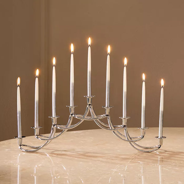 A Georg Jensen Harmony Menorah with nine lit white Hanukkah candles sits on a marble surface against a beige background.