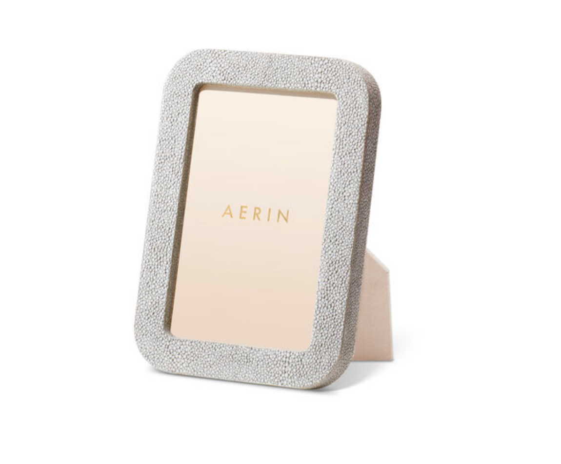 The Modern Shagreen Frame Dove by Aerin features a textured light gray embossed shagreen in a rectangular shape, standing upright on a white background, with "AERIN" in gold displayed on the placeholder inside the frame.