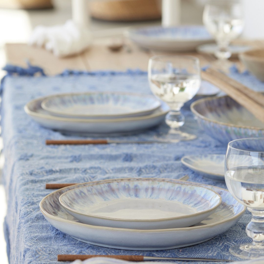 A close-up of a dining table featuring Costa Nova’s Brisa Blue Dinnerware, crafted in Portugal. Stoneware plates, water glasses, and wooden-handled cutlery create a cozy, inviting atmosphere.