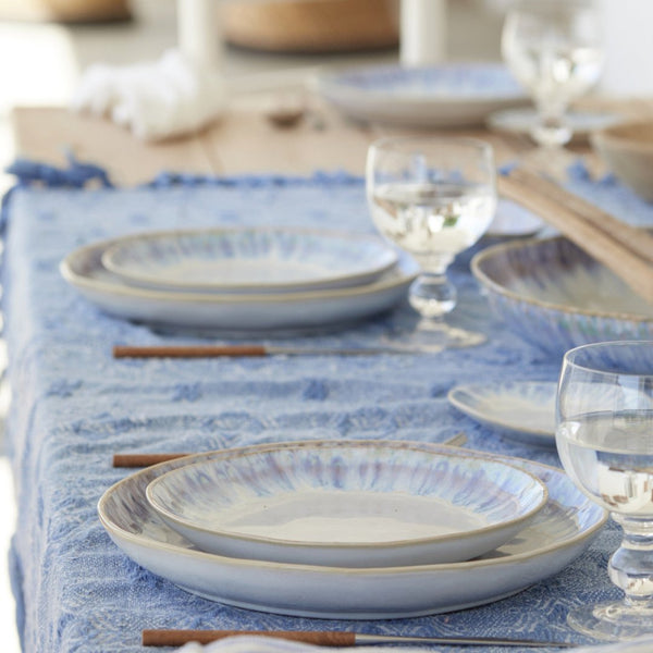 A close-up of a dining table featuring Costa Nova’s Brisa Blue Dinnerware, crafted in Portugal. Stoneware plates, water glasses, and wooden-handled cutlery create a cozy, inviting atmosphere.