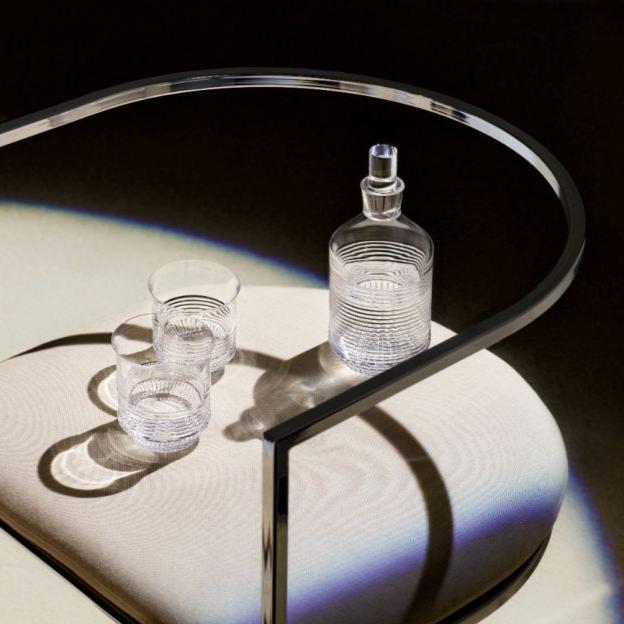 The Cadence Barware by Saint-Louis/Hermes, featuring a hand-blown crystal decanter and two linear-patterned tumblers, rests on a pale cushion beneath a modern curved metal chair, casting striking shadows and reflections.