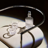 The Cadence Barware by Saint-Louis/Hermes, featuring a hand-blown crystal decanter and two linear-patterned tumblers, rests on a pale cushion beneath a modern curved metal chair, casting striking shadows and reflections.