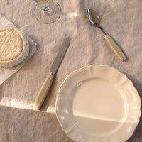 A cream-colored plate and Sabre Djembe Flatware 5 Piece Setting with faux horn handles rest on a beige tablecloth. Nearby are a glass of white wine and a wheel of soft cheese on parchment paper, with sunlight casting gentle shadows across the scene.