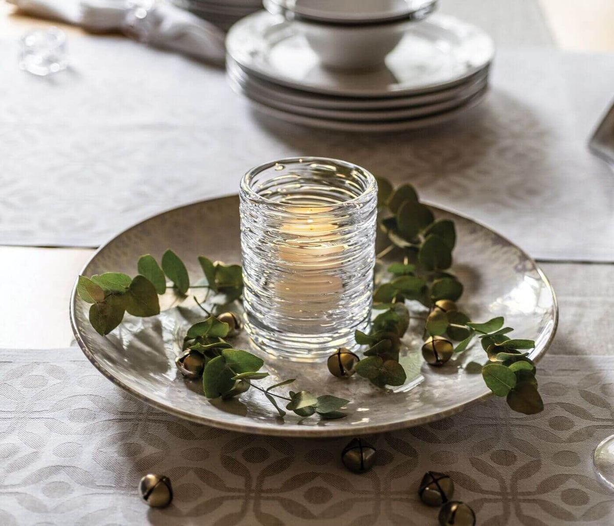 The Simon Pearce Echo Lake Tealight In Gift Box glows with a lit candle, displayed on a plate with green leaves and gold jingle bells, atop a patterned tablecloth with stacked plates and bowls in the background.
