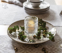 The Simon Pearce Echo Lake Tealight In Gift Box glows with a lit candle, displayed on a plate with green leaves and gold jingle bells, atop a patterned tablecloth with stacked plates and bowls in the background.