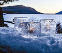 Three Simon Pearce Echo Lake Tealight candle holders in a gift box glow on snow at sunset, with a frozen lake and mountains in the background, creating a serene winter scene.