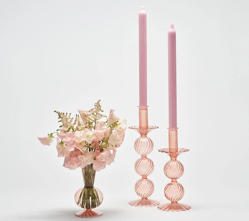 A Tess Bud Vase Set of 3 - Blush consisting of a small pink hand-blown glass vase filled with light pink flowers, next to two taller pink glass bud vases holding light pink taper candles.