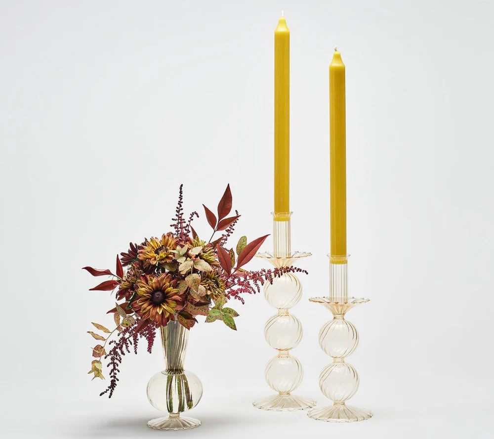 A small glass vase with autumn-colored flowers sits beside two tall yellow candles in Kim Seybert’s Iris Candlestick Holder Set of 2, beautifully arranged against a plain white background.