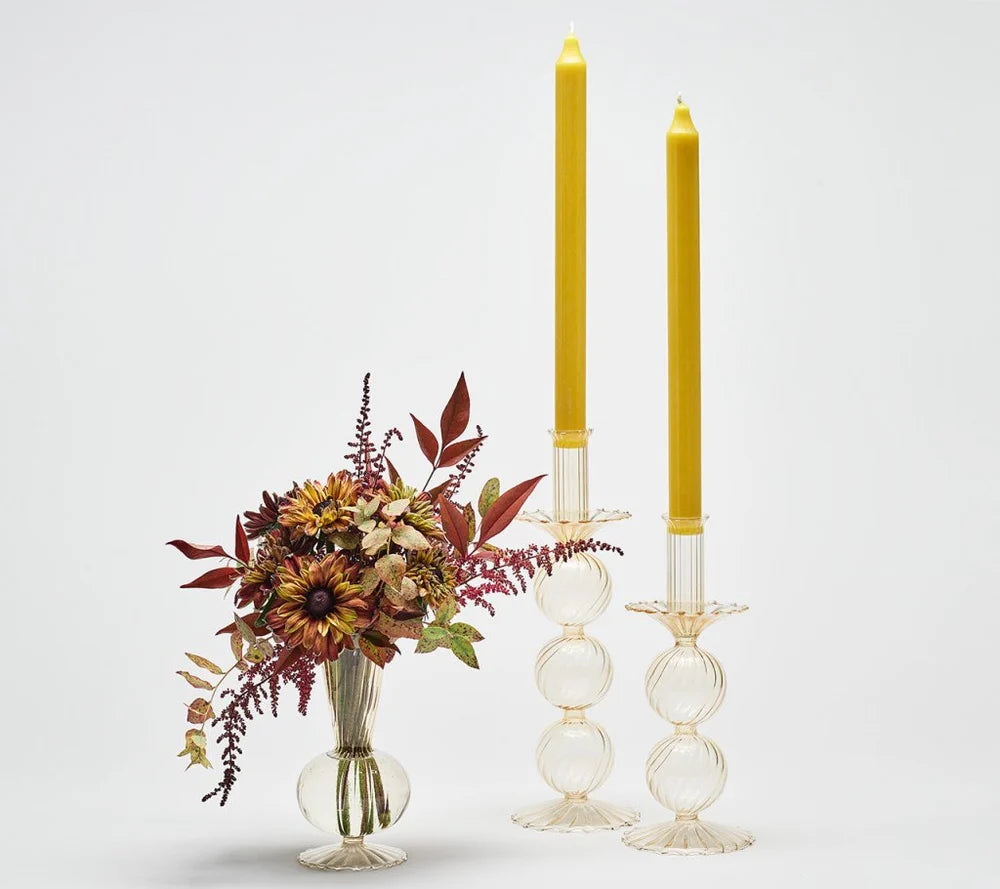 Kim Seybert’s Bella Candlestick Holder Set of 2, made from clear hand-blown glass and featuring tall yellow candles, stands beside a small glass vase filled with fall-colored flowers and foliage, all set against a plain white background.