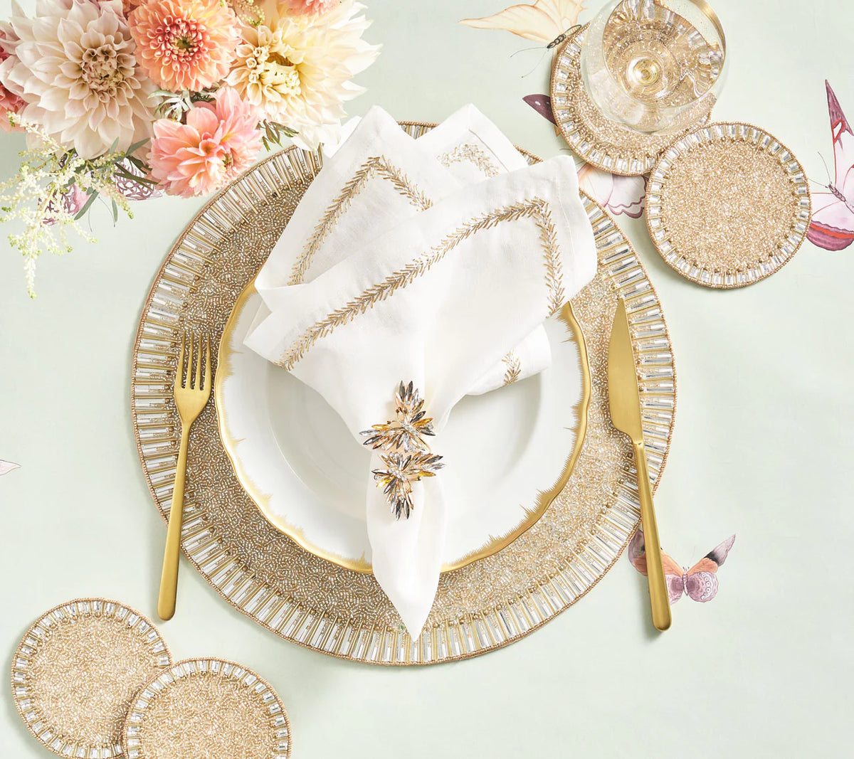 Elegant table setting with gold cutlery, white plate, glassware, beaded coasters, pink flowers, and a decorative napkin featuring the Kim Seybert Butterflies Napkin Ring Set of 4 on a pastel butterfly-patterned tablecloth.