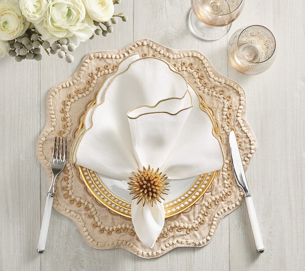 A sophisticated table setting featuring layered plates, a white napkin with gold trim secured by the Kim Seybert Stellar Napkin Ring Gold (Set of 4), complemented by white-handled fork and knife, two glasses, and white flowers in the background on a light wood surface.