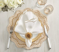 A sophisticated table setting featuring layered plates, a white napkin with gold trim secured by the Kim Seybert Stellar Napkin Ring Gold (Set of 4), complemented by white-handled fork and knife, two glasses, and white flowers in the background on a light wood surface.