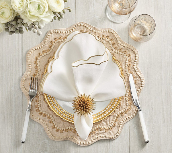 A sophisticated table setting featuring layered plates, a white napkin with gold trim secured by the Kim Seybert Stellar Napkin Ring Gold (Set of 4), complemented by white-handled fork and knife, two glasses, and white flowers in the background on a light wood surface.