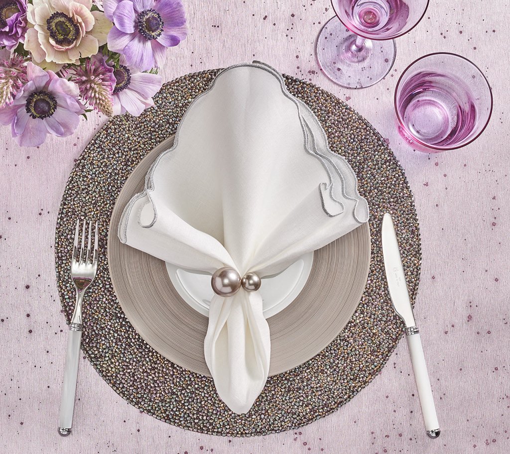 A table setting features a white scalloped napkin with the Kim Seybert Pearl Napkin Ring Grey & Silver Set of 4, atop layered plates on a beaded placemat. Fork and knife lie beside pink flowers and two pink glasses—perfect for wedding table decor.