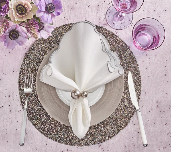 A table setting features a white scalloped napkin with the Kim Seybert Pearl Napkin Ring Grey & Silver Set of 4, atop layered plates on a beaded placemat. Fork and knife lie beside pink flowers and two pink glasses—perfect for wedding table decor.