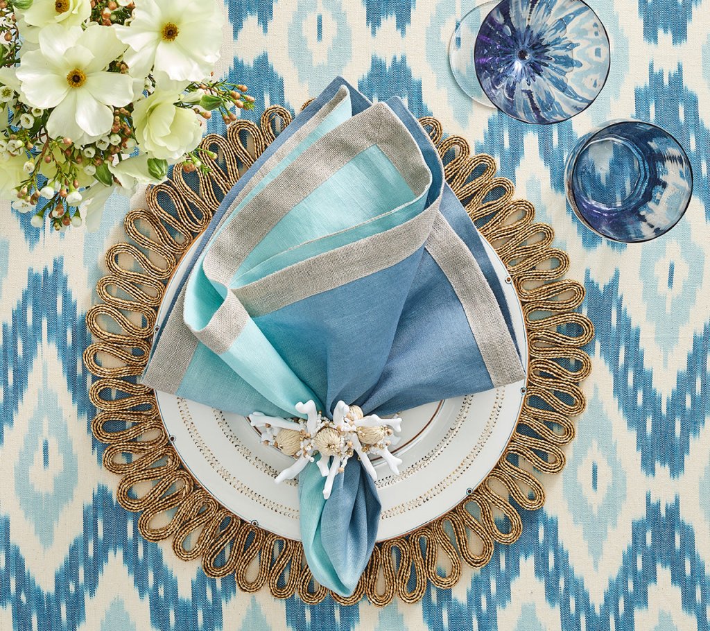 A Kim Seybert Dip Dye Napkins - Cool Tones napkin with a decorative ring rests on a gold-accented white plate and gold placemat, set on a blue and white patterned tablecloth with flowers and two blue glasses nearby.