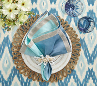 A Kim Seybert Dip Dye Napkins - Cool Tones napkin with a decorative ring rests on a gold-accented white plate and gold placemat, set on a blue and white patterned tablecloth with flowers and two blue glasses nearby.