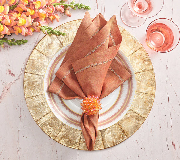 A table setting features the Kim Seybert Capiz Shell Placemat Champagne, a gold charger, white dinner plate, rust napkin with jeweled ring, two glasses of pink drink, and orange flowers on a light pink surface.