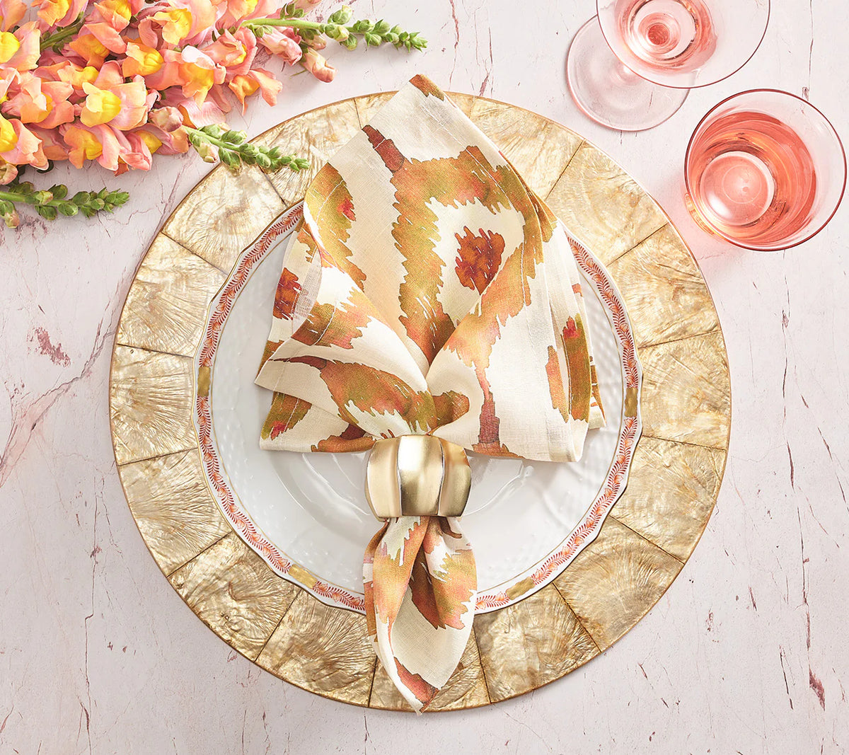 An elegant table is set with Kim Seybert Capiz Shell Placemat Champagne, gold-rimmed and white plates, a patterned napkin in a gold ring, two pink drinks, and orange flowers on a light pink marble surface.